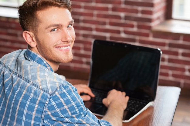 Jovem e criativo. vista traseira de um jovem bonito trabalhando em um laptop e olhando por cima do ombro enquanto está sentado em seu local de trabalho