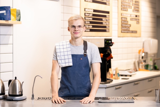 Jovem e feliz barista de sucesso em roupas de trabalho, aguardando no local de trabalho em um café enquanto espera por novos convidados