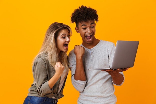 Jovem e feliz casal apaixonado, isolado na parede amarela, usando um laptop, fazendo um gesto de vencedor