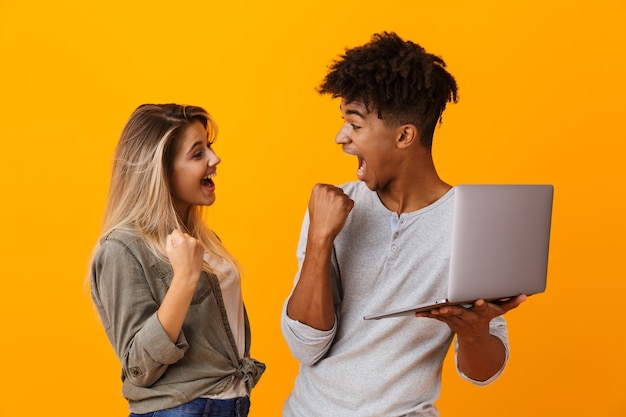 Jovem e feliz casal apaixonado, isolado na parede amarela, usando um laptop, fazendo um gesto de vencedor