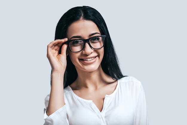 Jovem e inteligente. mulher jovem e atraente de camisa branca e óculos sorrindo em pé contra um fundo cinza