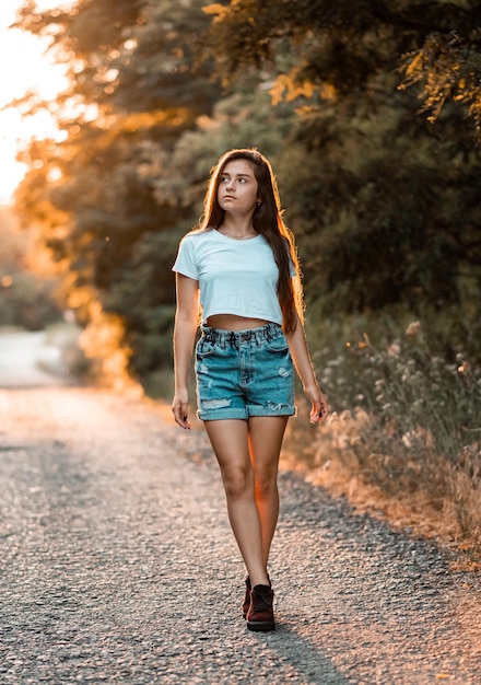 Jovem e linda adolescente morena em camiseta branca e roupas de shorts azuis posando no parque pôr do sol