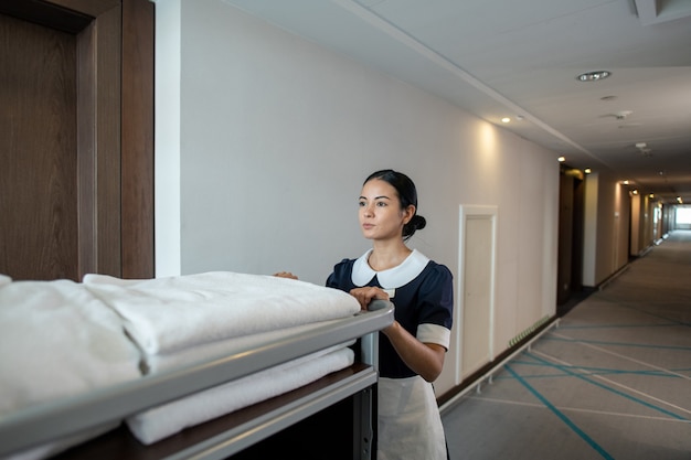 Foto jovem e linda morena camareira ou trabalhadora de hotel empurrando carrinho com toalhas limpas dobradas e outras coisas enquanto se move pelo corredor