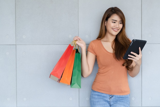 Foto jovem e linda mulher asiática feliz com carinha está usando smartphone ou tablet e carregando sacolas de compras