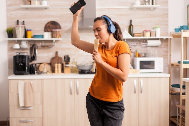 Jovem e linda mulher dançando enquanto ouve música com fones de ouvido azuis sem fio na cozinha