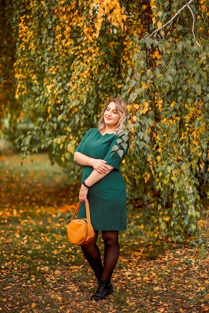Jovem e linda mulher de tamanho grande em um vestido verde fica perto de uma árvore de outono