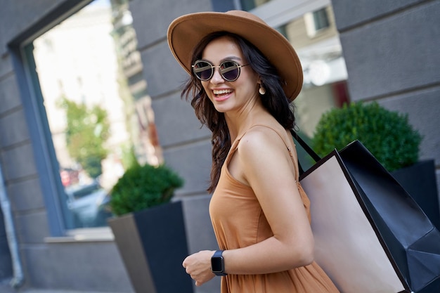 Foto jovem e linda mulher elegante usando chapéu de óculos escuros e vestido de verão com sacola de compras, olhando para