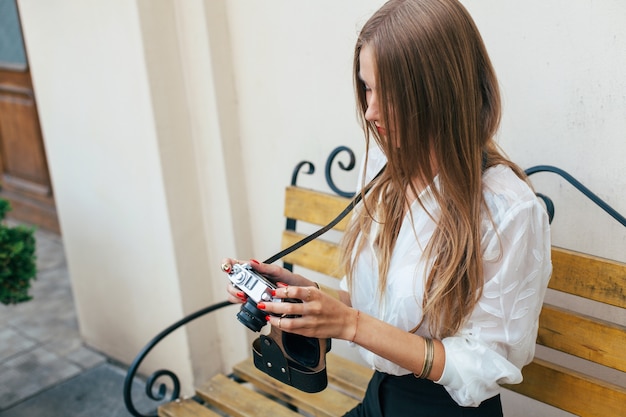 Jovem e linda mulher está olhando fotos com sua câmera sentada no banco no verão