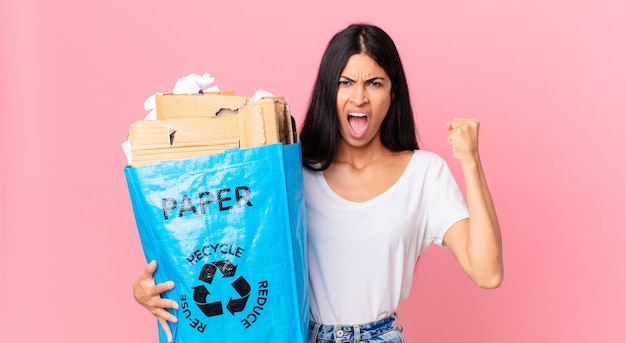 Foto jovem e linda mulher hispânica gritando agressivamente com uma expressão de raiva e segurando um saco de papel para reciclar