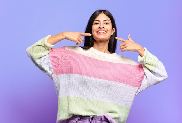 Jovem e linda mulher hispânica sorrindo com confiança apontando para o próprio sorriso largo, atitude positiva, relaxada e satisfeita