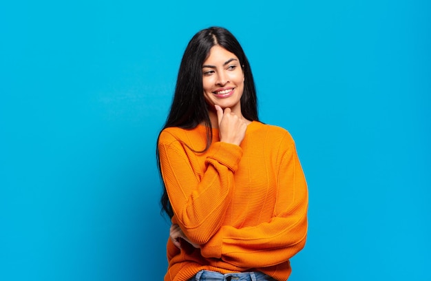 Jovem e linda mulher hispânica sorrindo com uma expressão feliz e confiante com a mão no queixo, pensando e olhando para o lado