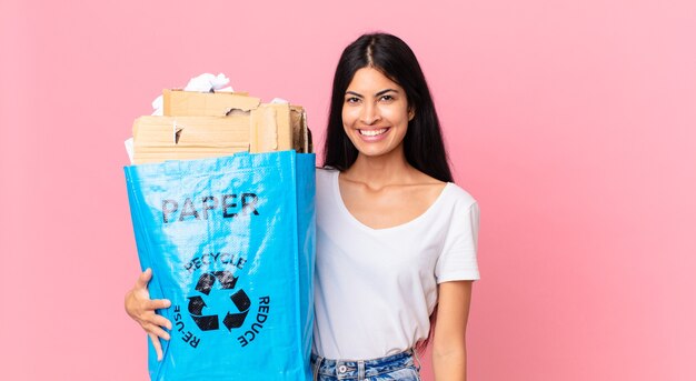 Jovem e linda mulher hispânica sorrindo feliz com uma mão no quadril e confiante, segurando um saco de papel para reciclar