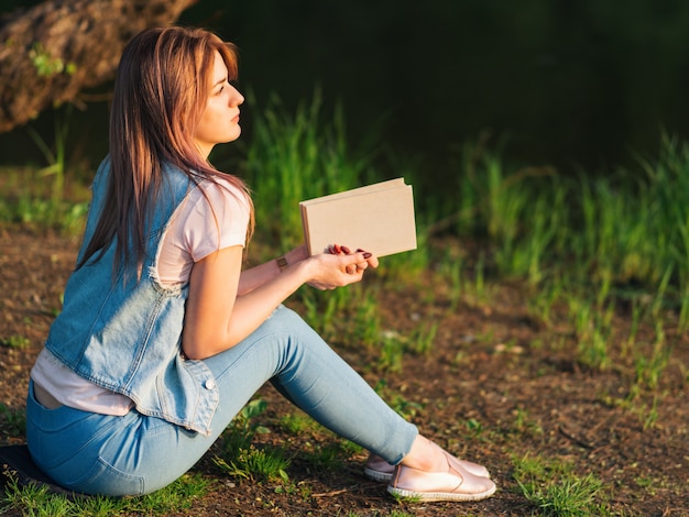 Jovem e linda mulher lendo um livro sobre a margem do rio durante o pôr do sol