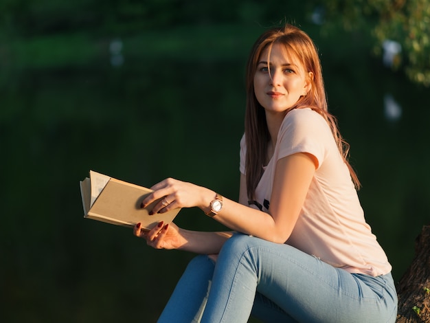 Jovem e linda mulher lendo um livro sobre a margem do rio durante o pôr do sol