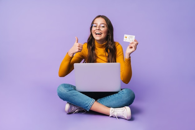 Foto jovem e linda mulher segurando um cartão de crédito e usando o laptop enquanto está sentado com as pernas cruzadas isoladas