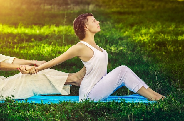 Jovem e mulher vestida com uma túnica branca, fazendo massagem tailandesa com exercícios de ioga, sentado no tapete. Parque de verão ensolarado com gramado verde ao fundo