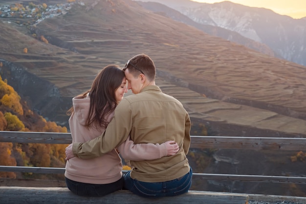 Jovem e namorada morena sentados em um banco em um mirante contra pitorescas montanhas antigas ao pôr do sol