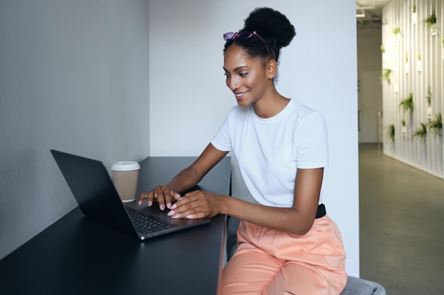 Jovem e sorridente mulher afro-americana casual trabalhando em laptop no espaço de co-trabalho moderno