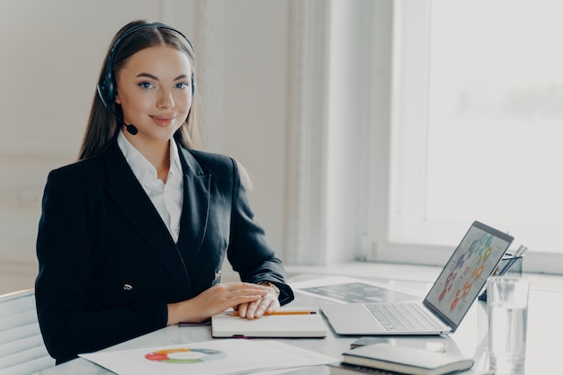 Jovem economista bem-sucedida em terno formal e fone de ouvido sorrindo para a câmera, analisando dados financeiros no laptop, empresária sentada em um escritório moderno e sentindo-se satisfeita com os resultados do projeto