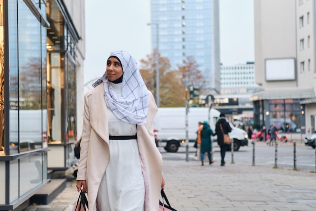 Jovem elegante árabe em hijab sonhadoramente andando pela rua da cidade com sacolas de compras