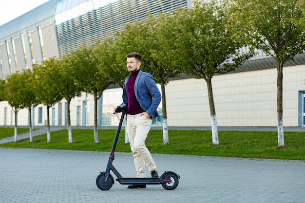 Jovem elegante bonito com e-scooter em pé na calçada perto do aeroporto na sunset