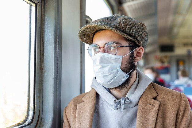 Jovem elegante com barba, óculos e uma máscara médica em um trem olha pela janela. regime de auto-isolamento e distância social durante a pandemia de coronavírus. fechar-se. medidas de precaução.