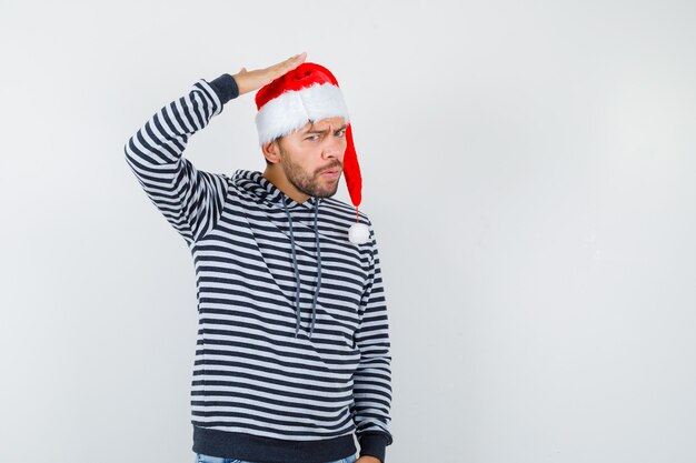 Jovem elegante com capuz, chapéu de Papai Noel segurando a mão na cabeça e parecendo pensativo.