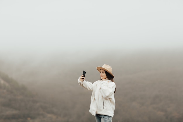 Jovem elegante com chapéu, tome uma selfie no fundo das belas montanhas de nevoeiro pela manhã.