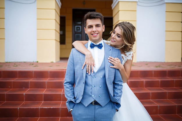 Jovem elegante com o traje do noivo e da noiva linda garota em um vestido branco com um trem andando no fundo de uma grande casa com colunas no dia do casamento