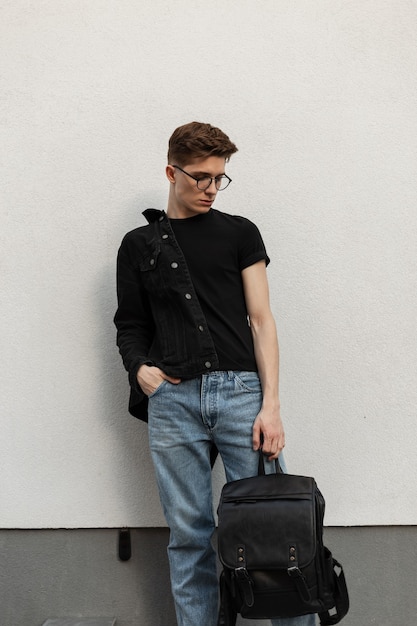 Jovem elegante com penteado em jaqueta jeans preta em jeans com óculos com mochila de couro fica e olha para baixo perto de um edifício vintage na cidade. cara urbano. moda casual americana.