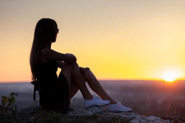 Jovem elegante de vestido curto preto e sapatos de tênis branco sentado em uma rocha relaxando ao ar livre na noite de verão Senhora elegante apreciando o pôr do sol quente na natureza