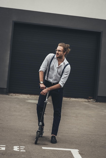 Jovem elegante e bonito com roupa formal e óculos andando de scooter no estacionamento