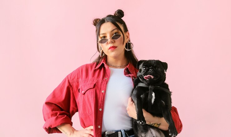 Foto jovem elegante e incomum posando segurando um cachorrinho pug preto nos braços na superfície rosa