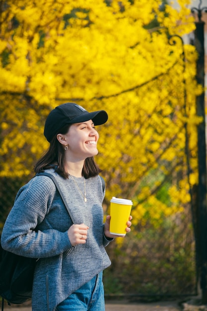 Jovem elegante em cap bebendo café ao ar livre de um copo de papel amarelo arbustos florescendo amarelos no fundo