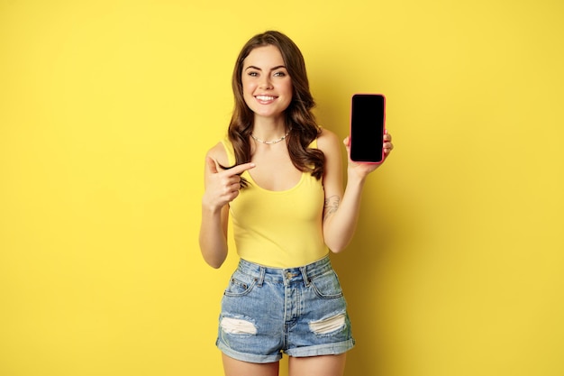 Foto jovem elegante mostrando a tela do celular, apontando para a tela do smartphone, interface do aplicativo ou loja online, sorrindo satisfeito, de pé sobre fundo amarelo