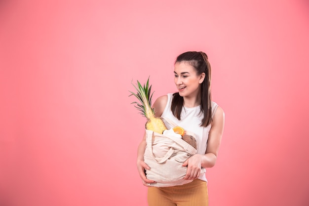 Jovem elegante sorrindo e segurando um saco ecológico com frutas exóticas em um espaço de cópia de parede rosa.