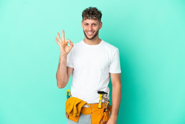 Jovem eletricista loiro isolado em fundo verde mostrando sinal de ok com os dedos