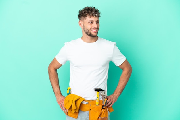 Jovem eletricista loiro isolado em fundo verde posando com braços no quadril e sorrindo