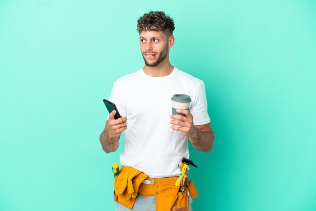 Jovem eletricista loiro isolado em fundo verde segurando café para levar e um celular enquanto pensava em algo