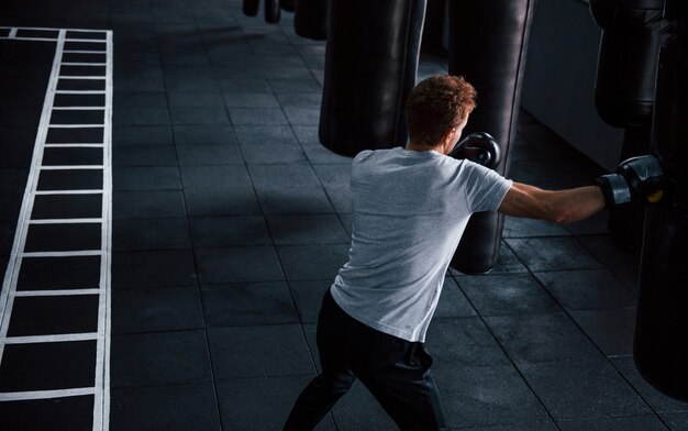 Jovem em camisa branca e luvas protetoras de boxe, fazendo exercícios no ginásio com empurrar o saco.