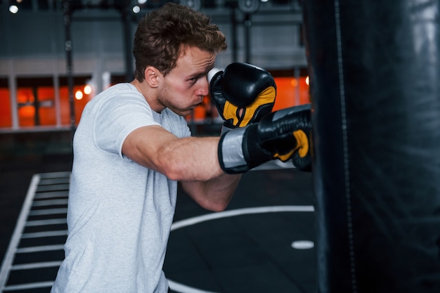 Jovem em camisa branca e luvas protetoras de boxe, fazendo exercícios no ginásio com empurrar o saco.