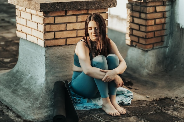 Jovem em depressão sente-se com os joelhos abraçados perdidos em maus pensamentos pessimistas e preparando-se para treinar em um terraço na cobertura.