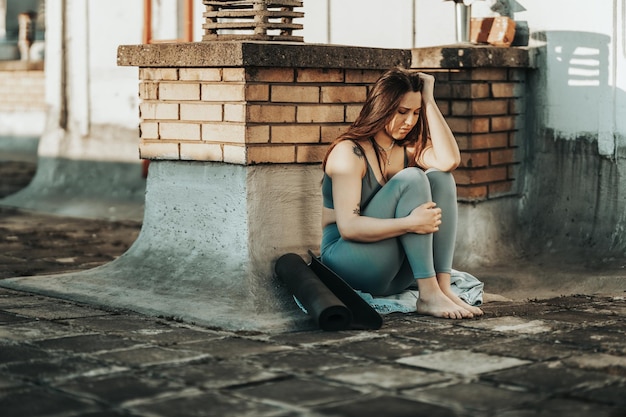 Jovem em depressão sente-se com os joelhos abraçados perdidos em maus pensamentos pessimistas e preparando-se para treinar em um terraço na cobertura.