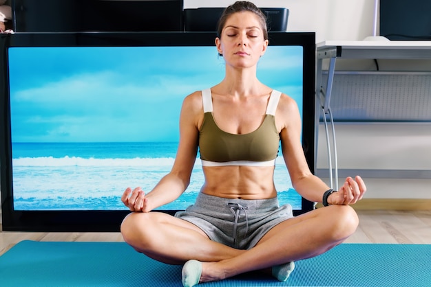 Jovem em forma de mulher fazendo ioga asana com os olhos fechados dentro de casa, perto da tela da TV, isolada em sua casa