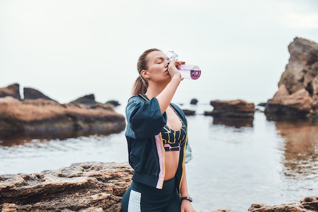 jovem em roupas esportivas bebendo água enquanto está sentado na pedra na praia.