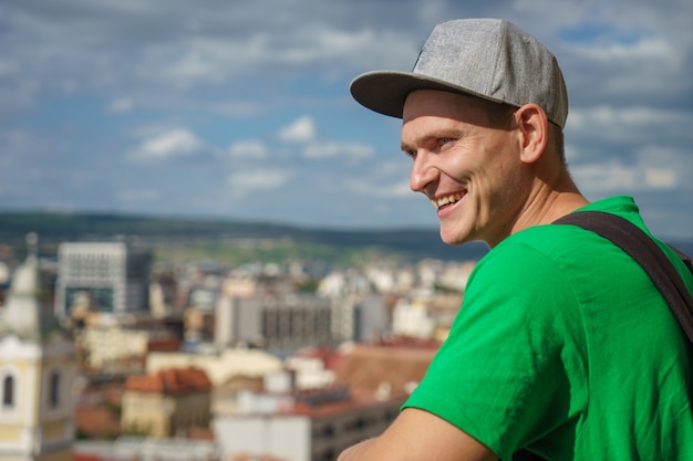 Jovem em um boné de beisebol cinza e camiseta verde no fundo da cidade e o céu azul com nuvens.