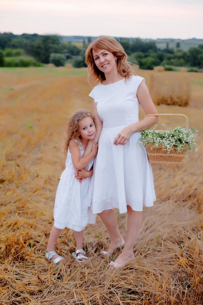 jovem em um vestido branco com uma cesta de flores está de pé no campo abraçando sua filha