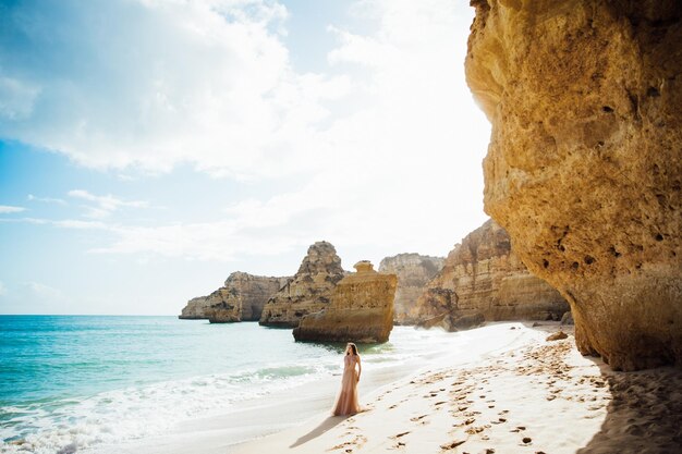 Jovem em um vestido longo andando na praia perto do mar.