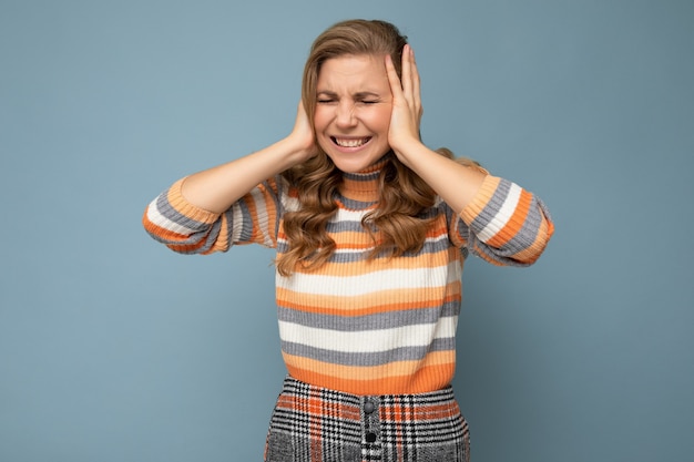 Jovem emocional linda loira encaracolada mulher com emoções sinceras, vestindo suéter listrado casual, isolado em um fundo azul com espaço de cópia e cobrindo as orelhas, tentando não ouvir.