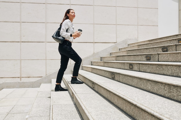 Jovem empreendedora séria com café para viagem subindo as escadas ao correr para o trabalho pela manhã